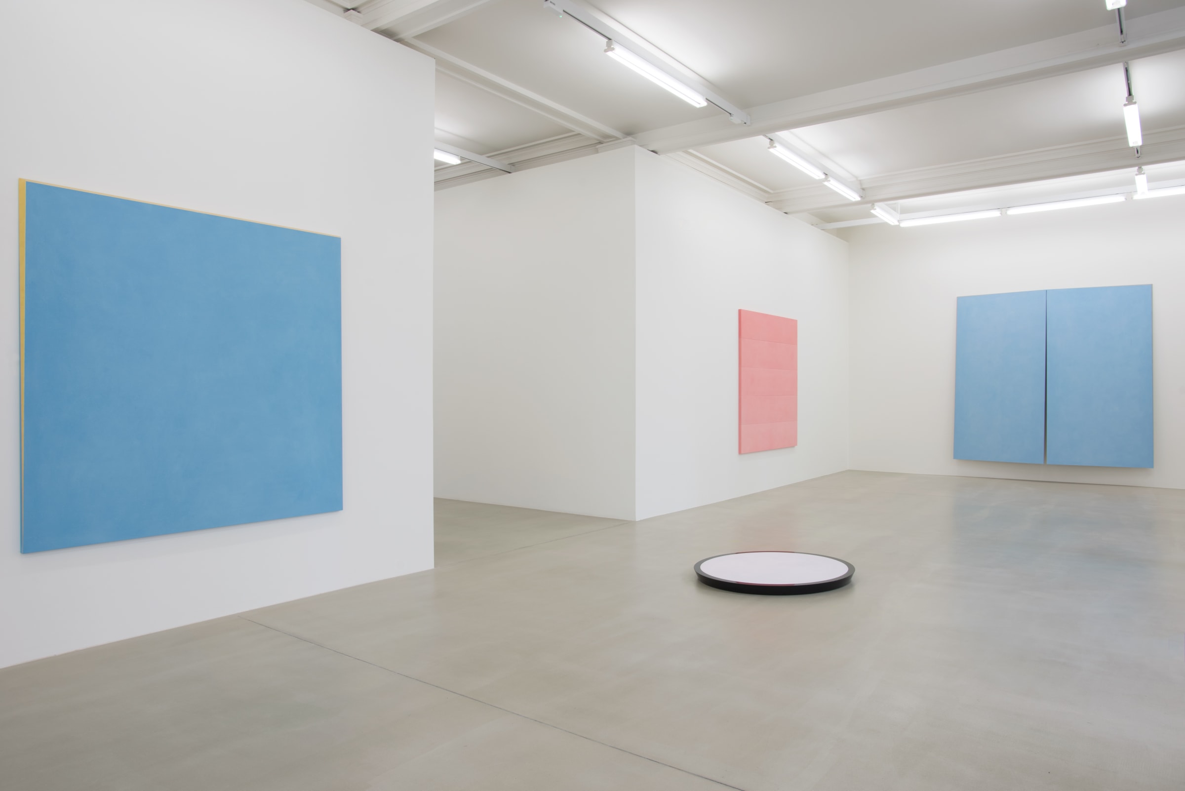 In a large white space with grey stone floors, a white circular sculpture with black trimming lies on the floor. On the walls surrounding it hang a light blue painting with gold trim, a light pink painting, and a larger blue painting, bisected.