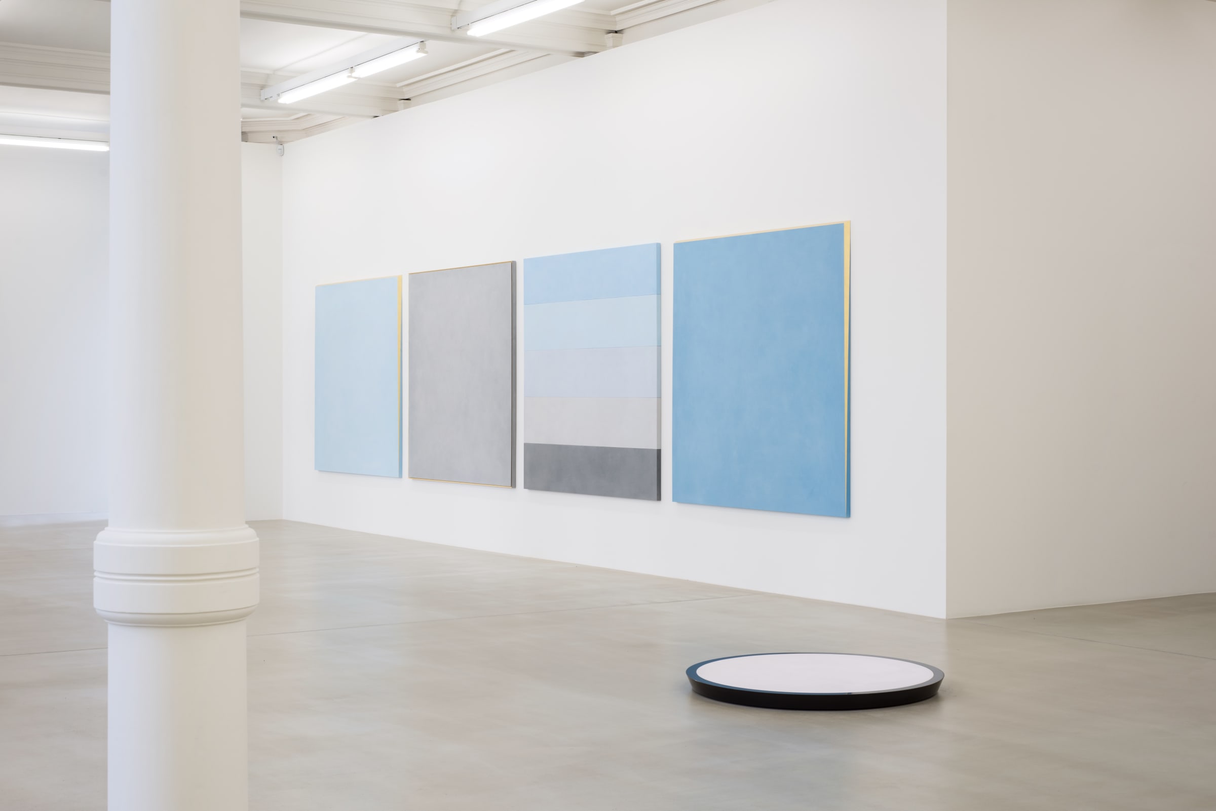 Four paintings hang on a white wall. From the left: solid light blue with gold framing, solid light gray with gold framing, 5 strips: light blue, lighter blue, light grey, grey,  darker grey, and finally, sky blue with gold framing.
