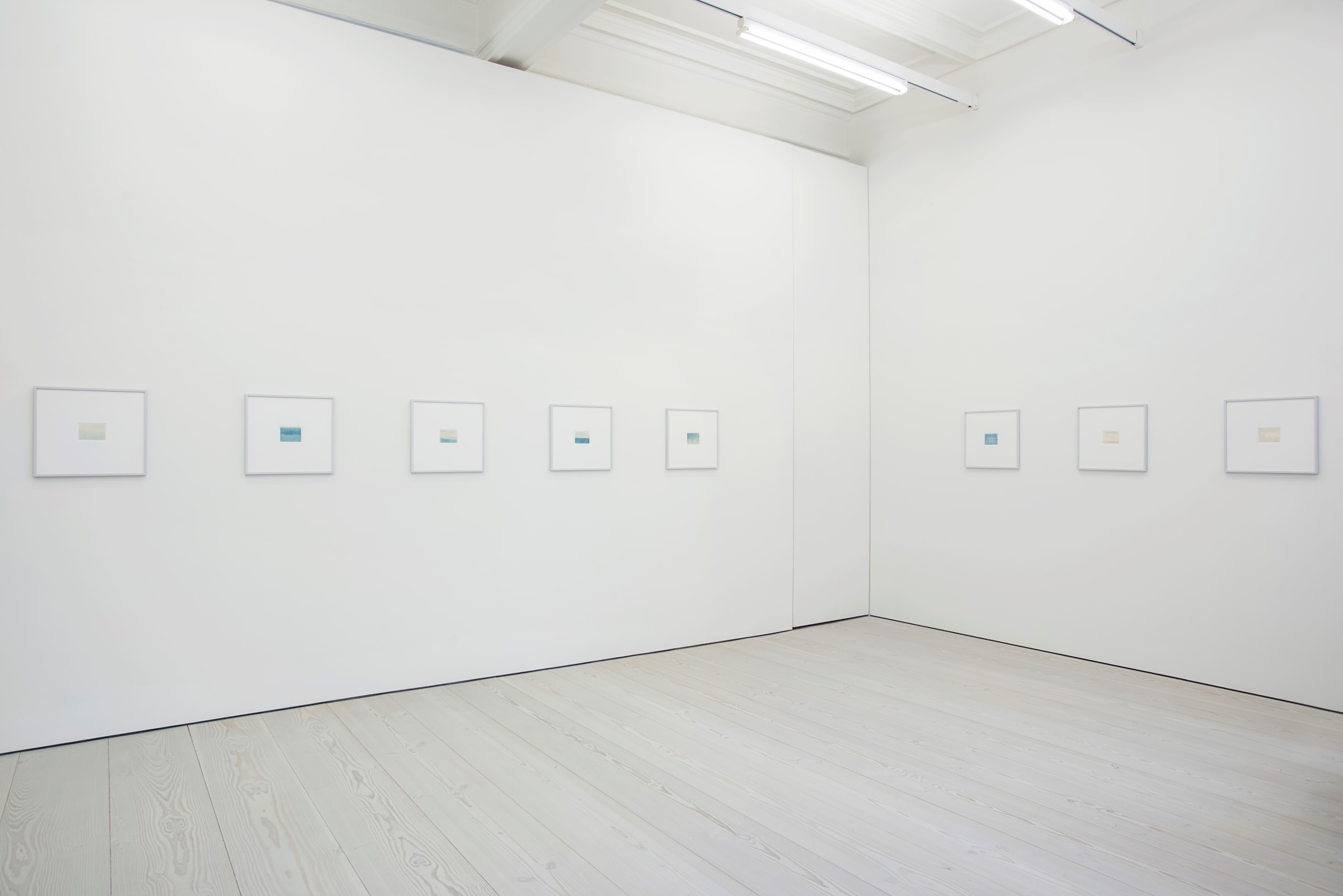 In a white space, 8 small drawings, 5 on the left wall and 3 on the right, hang. They are framed in white, and mostly made up of blues and yellows, like sunsets.