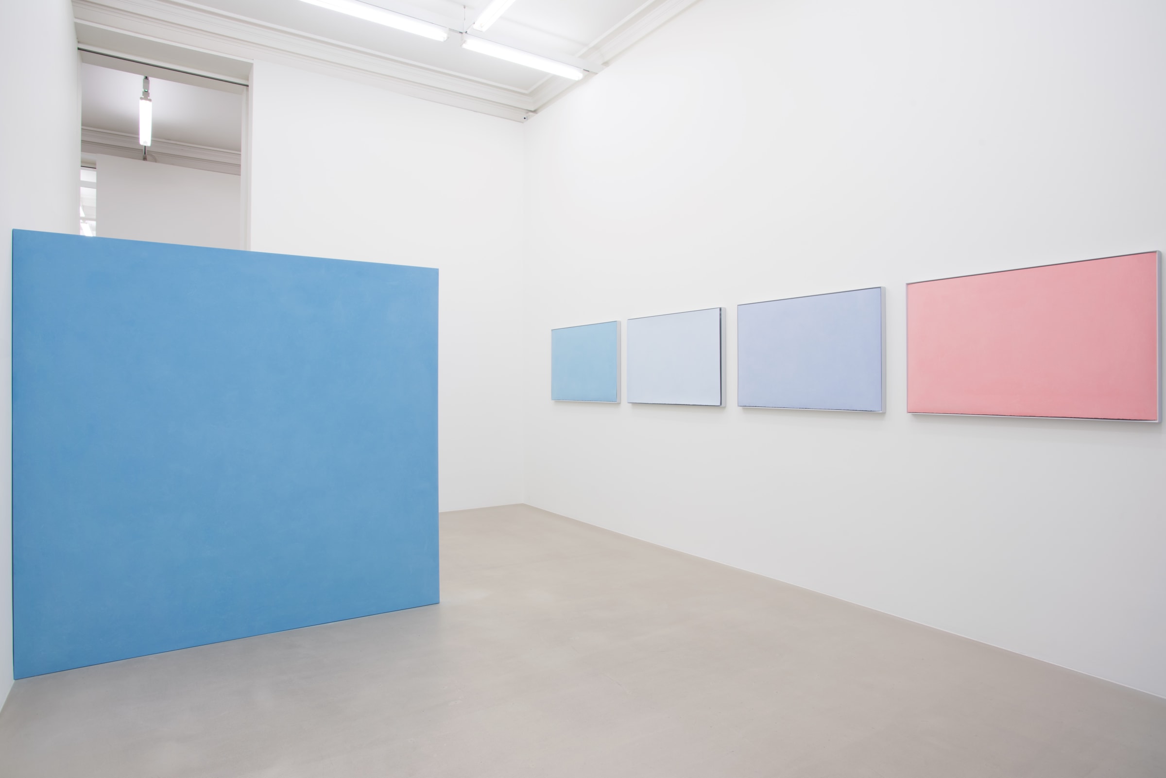 4 pastel colored paintings hang horizontally on the wall on the right. A large, light blue floating wall comes out perpendicularly from the wall on the left. 
