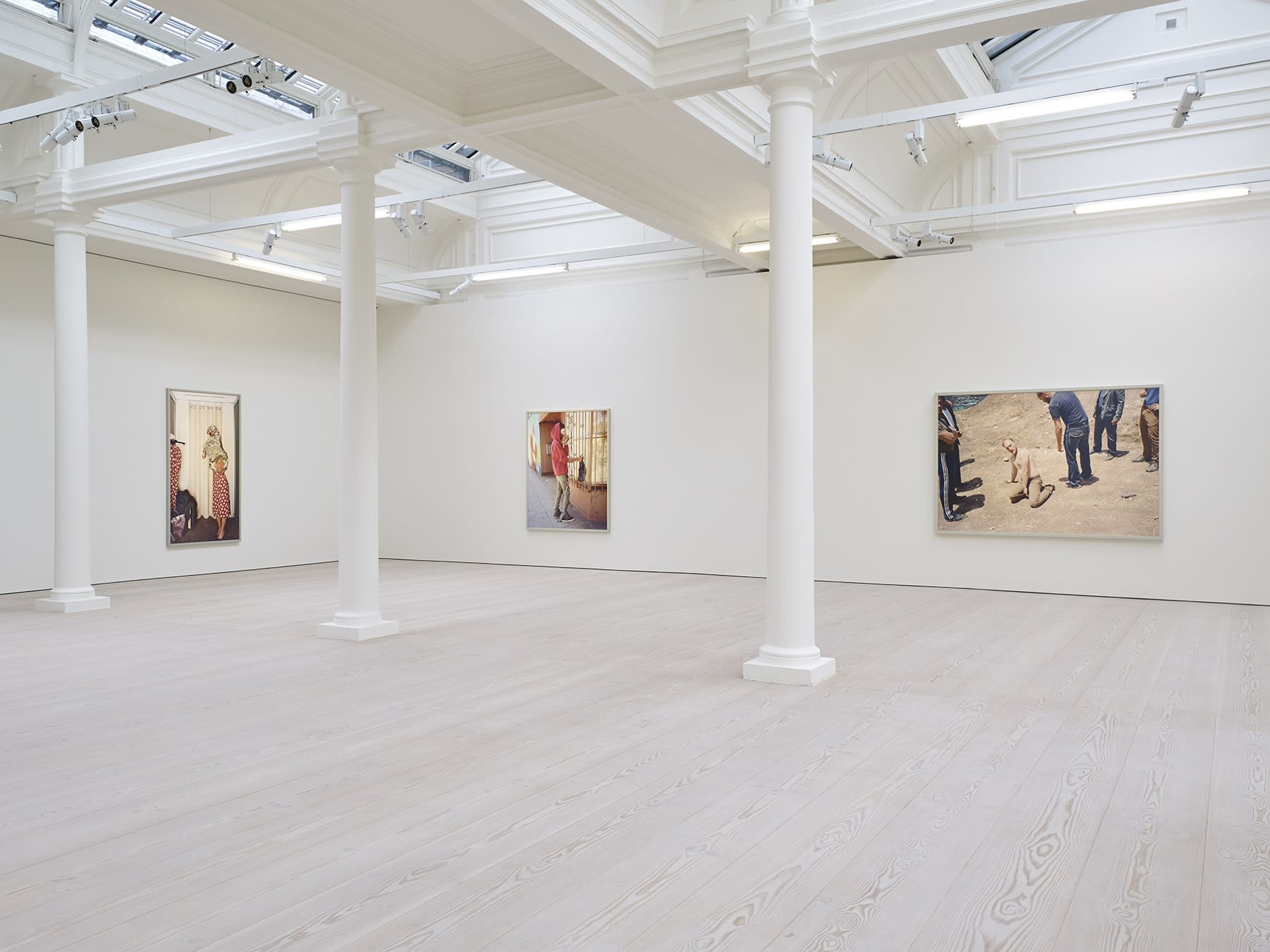 3 large photographs hang in a white room with columns and skylights. From left to right: A figure in a red polka dot dress; a man in a red sweatshirt and white mask; and a shirtless man on the ground surrounded by 6 clothed men. 