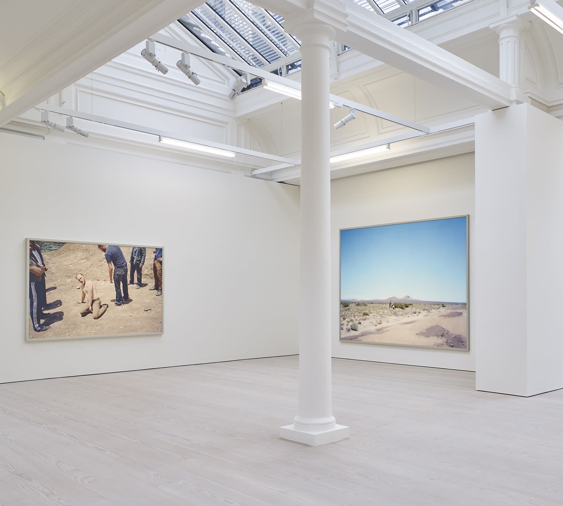 2 large photographs hang in the corner of a white room with skylights. On the left a shirtless man on the ground surrounded by 6 clothed men, and on the right a desert landscape. 
