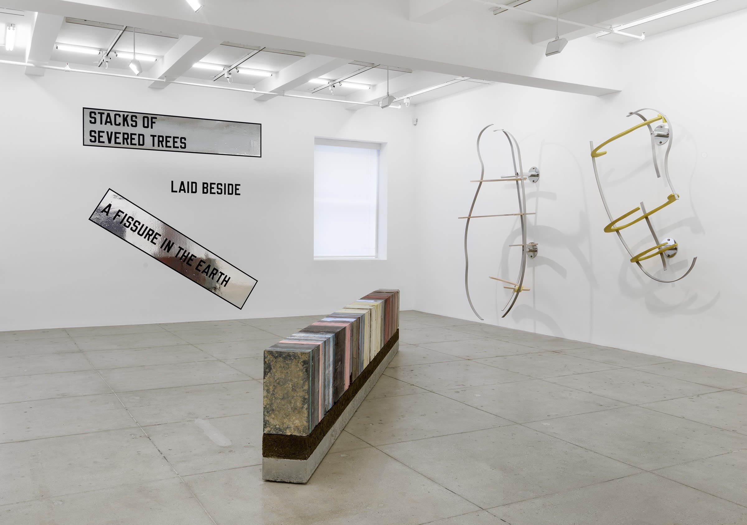 Various artworks are arranged in the corner of a white room with a window; text on a reflective banner, metal armatures painted white and yellow, and a long, rectangular multi-colored sculpture. 