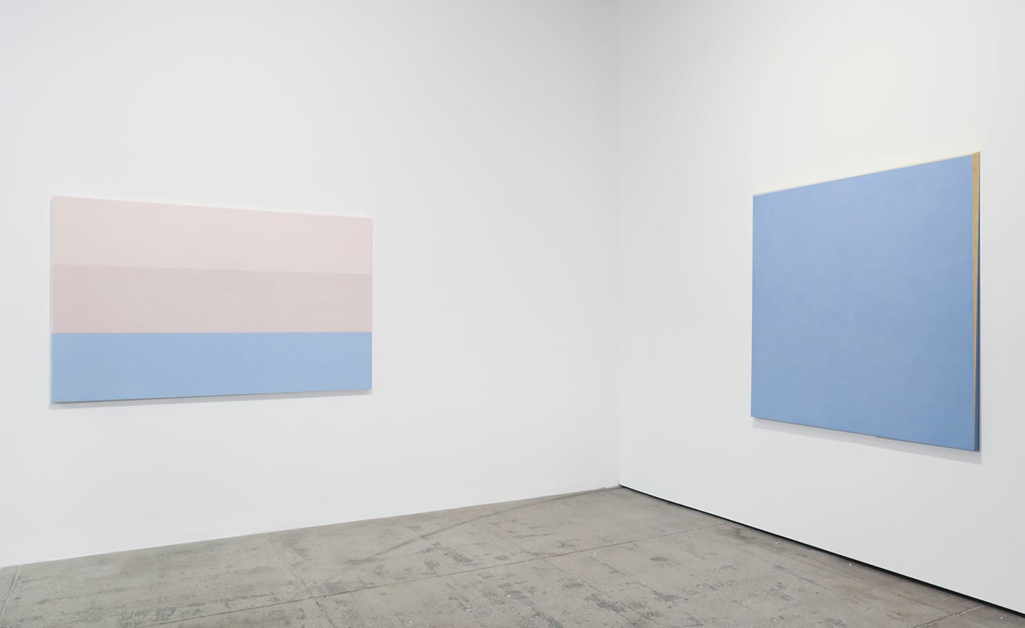 2 pastel colored paintings in pink and blue hang in the corner of a white room. 