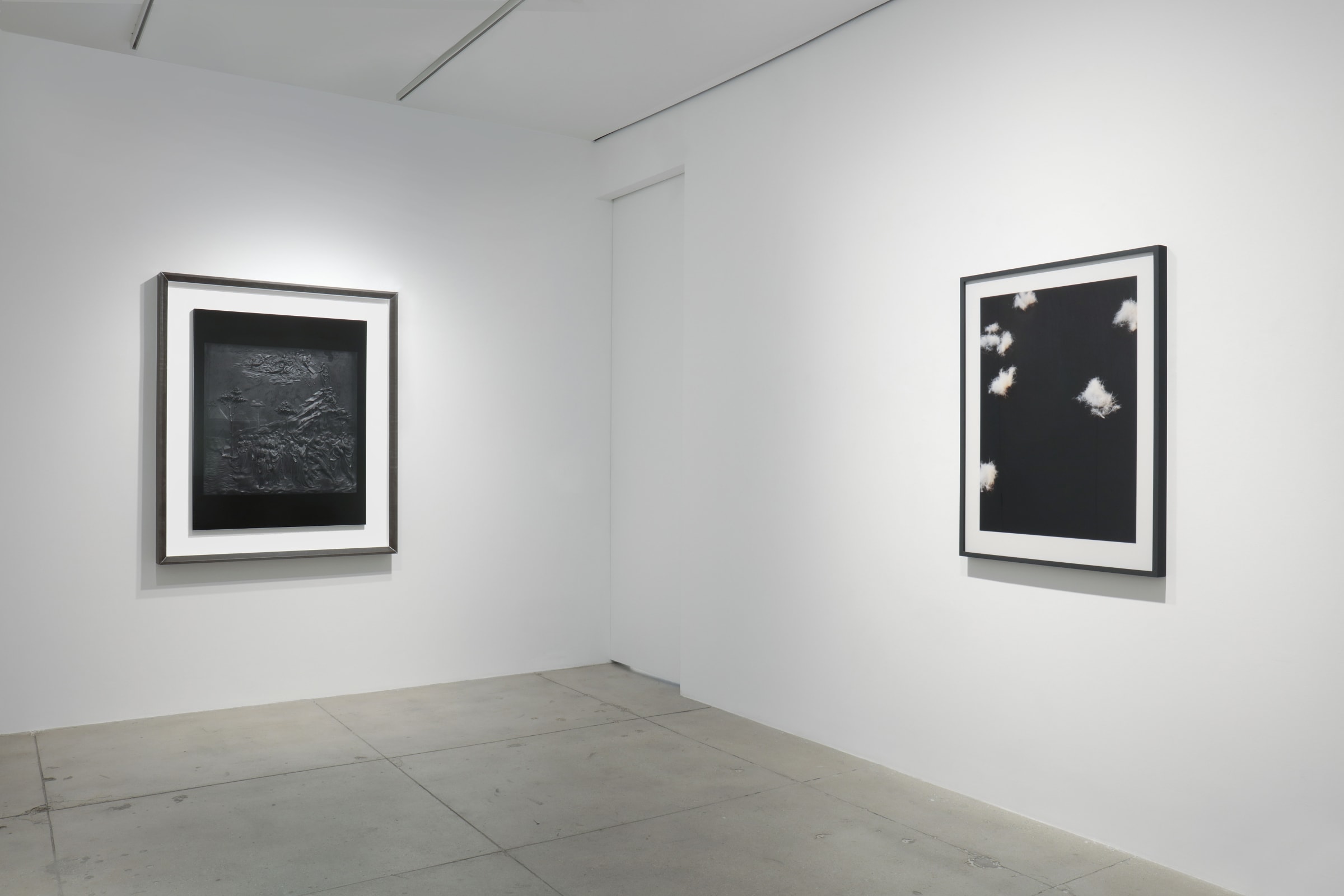 2 framed artworks hang in the corner of a white room. On the left appears to be an embossed landscape, while on the right is a black and white photograph of clouds. 