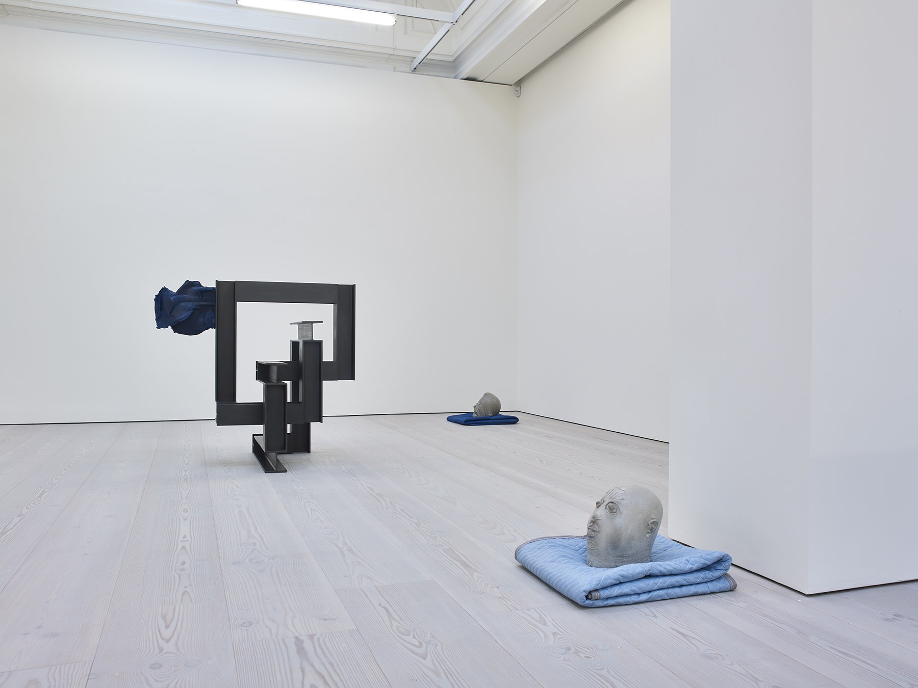 Gallery view, two abstract clay heads rest on blue blankets. To the left there is a geometric black square sculpture.