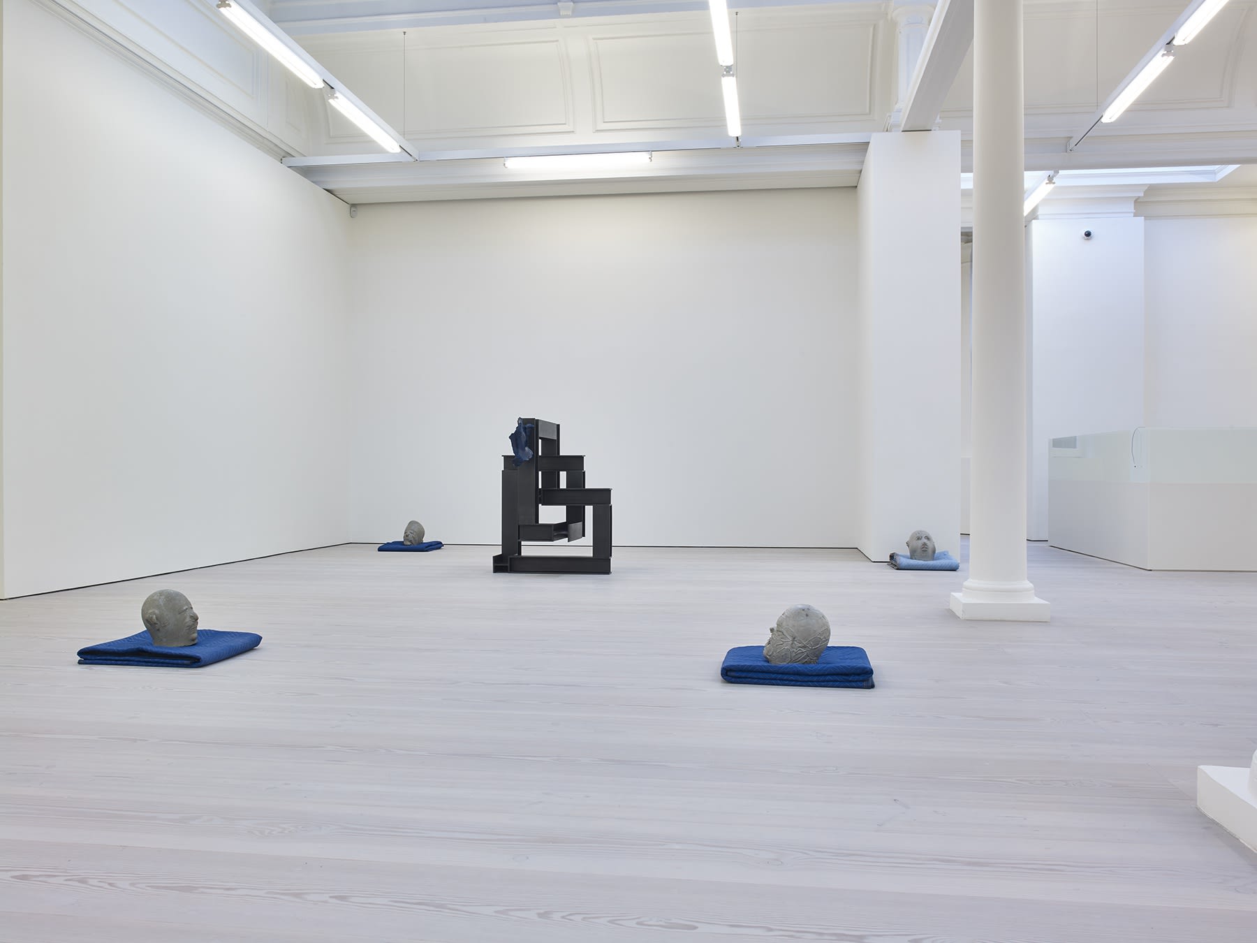 Gallery view, abstract clay heads rest on blue blankets, there is one geometric black square sculpture.