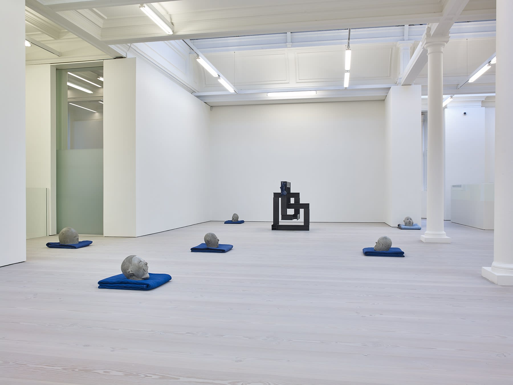 Gallery view, abstract clay heads rest on blue blankets, there is one geometric black square sculpture.