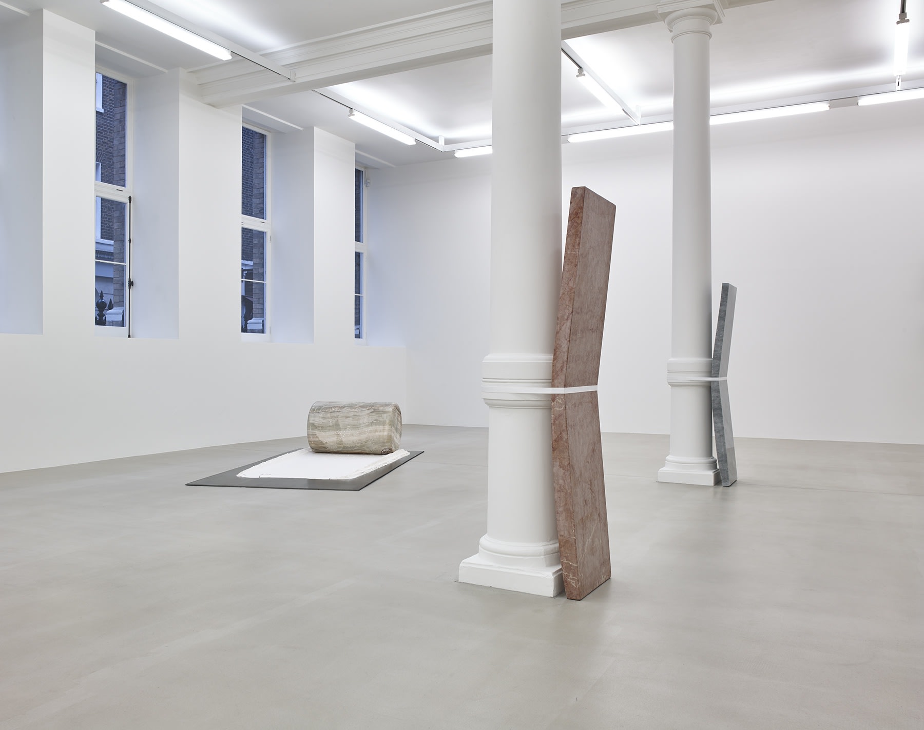 Installation view, slabs of stone strapped to pillars in a row and a cylinder stone on pallet. 