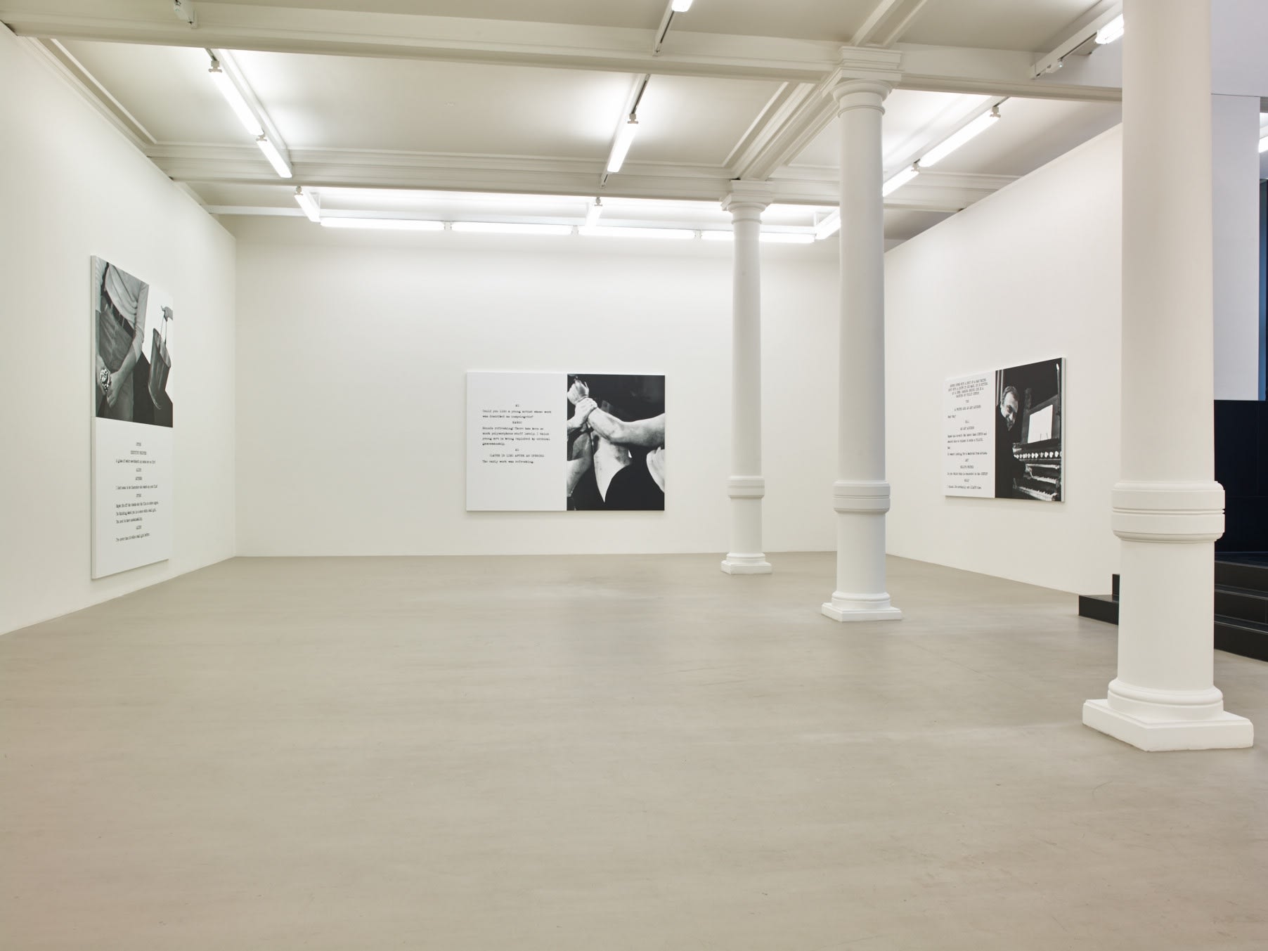 In a large white space with columns and a long skylight, 3 large paintings hang. Each are roughly half image and half text, which is in the format of a film script.