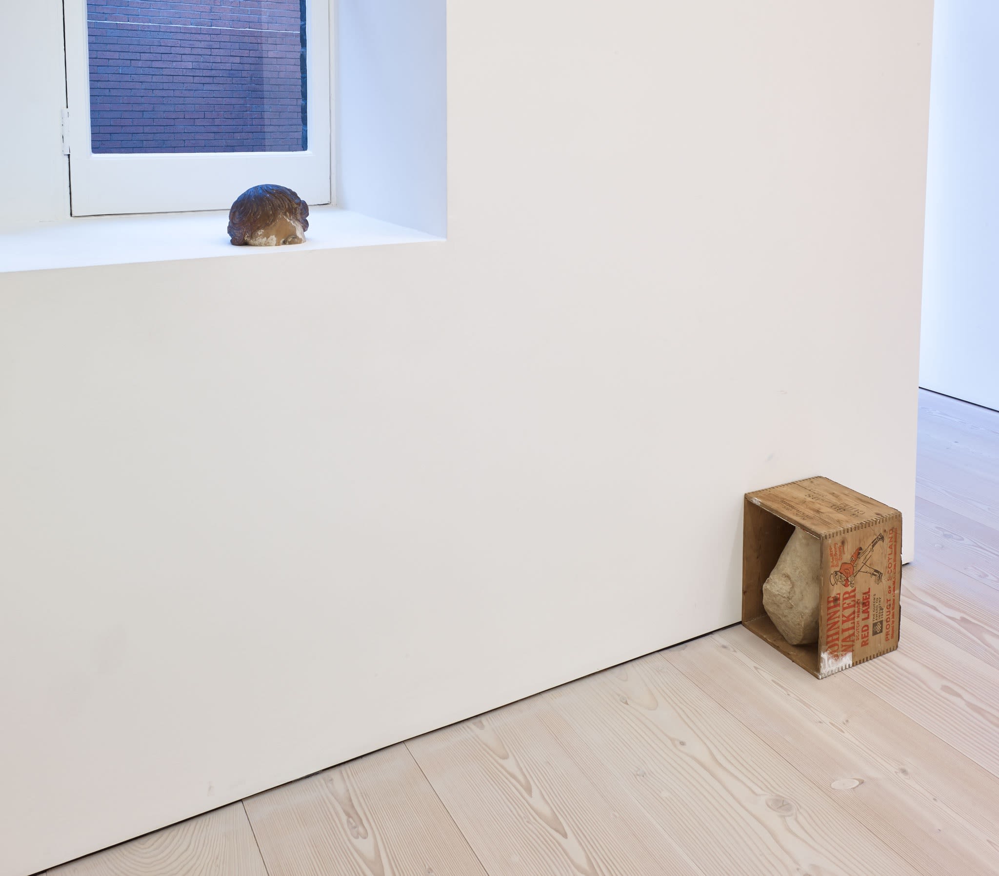 A small, unidentified object sits on a deep, white windowsill. On the white wooden floor, another sculpture sits: a large rock, stuck inside a sideways wooden box, which reads JOHNNIE WALKER RED LABEL.