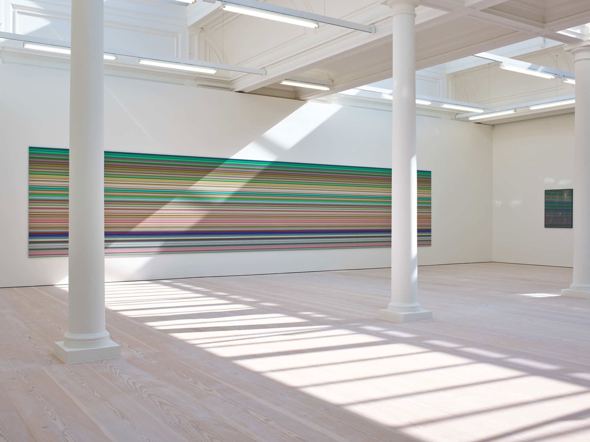 In a large white space with columns and skylights, an enormous painting hangs, light from the skylight shining on it. It is made up of hundreds of perfect horizontal strips of various colors, mostly light green in hue, with a great deal of reds and blues.