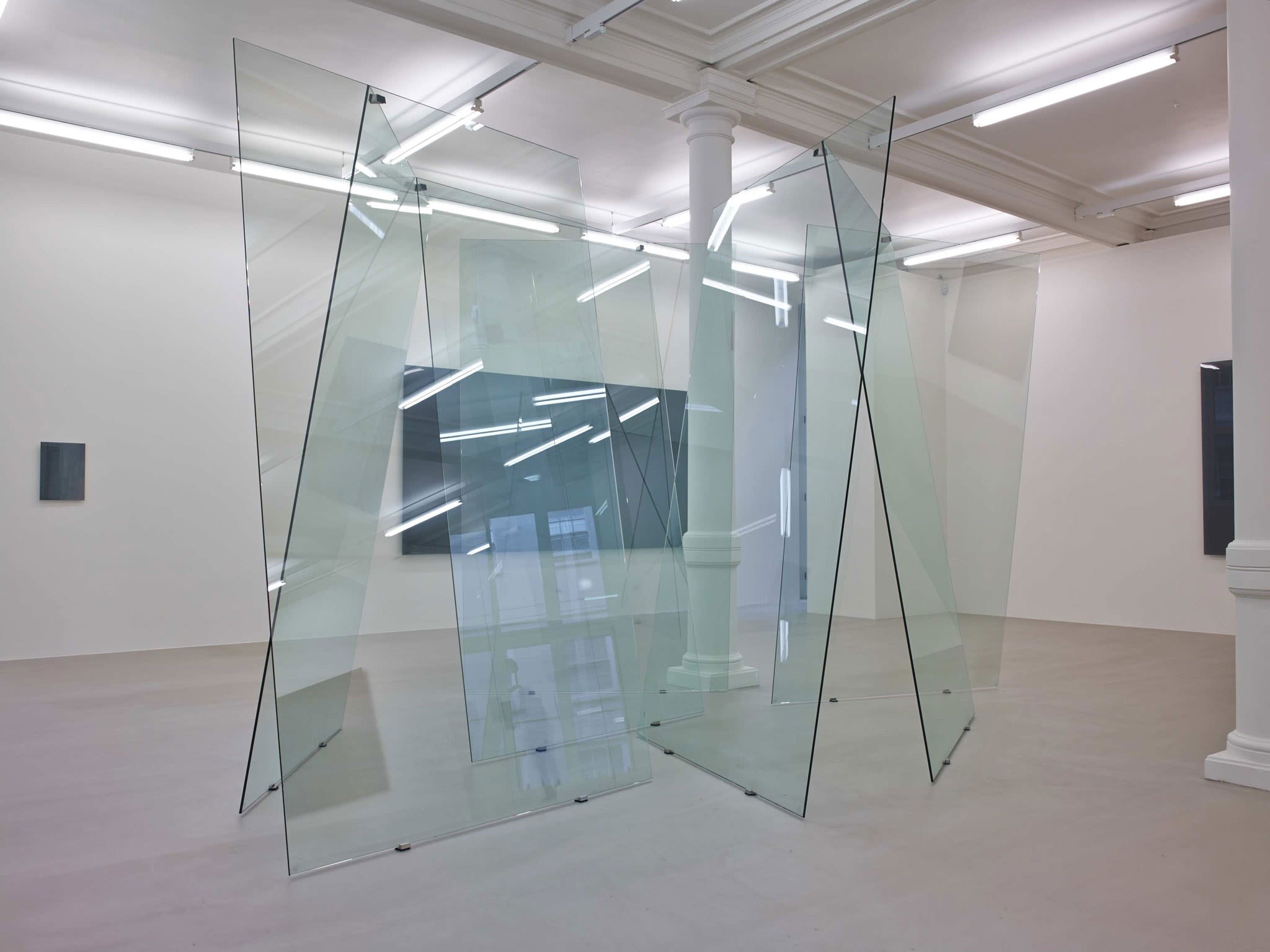 In a large white space with columns and many windows, several glass planes lean on each other unevenly, making a large sculpture.