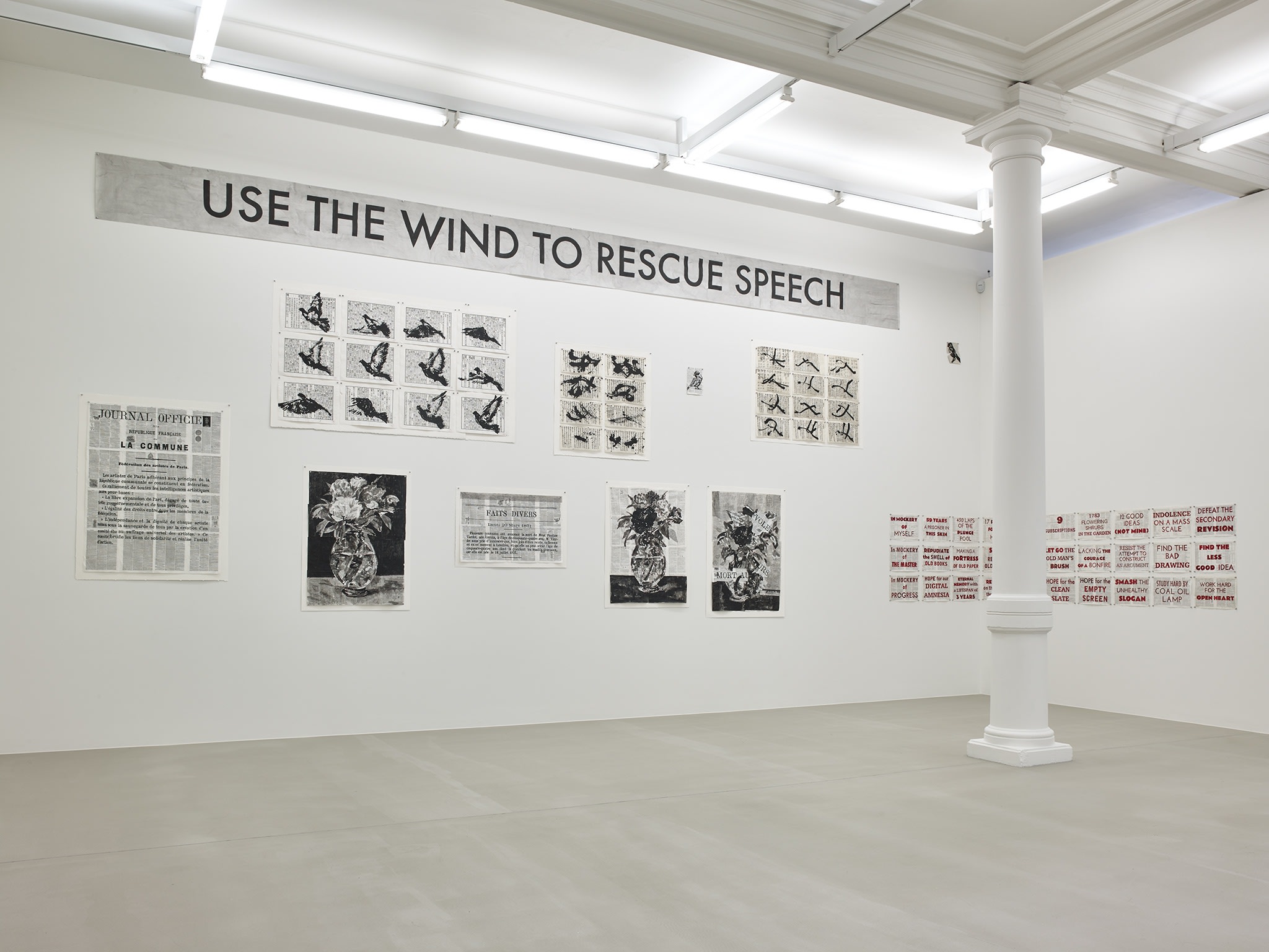 In a large white space with columns, many black and white paintings hang, of various sizes, all made up of smaller pieces of paper, except a large sign which hangs overhead, reading: USE THE WIND TO RESCUE SPEECH.