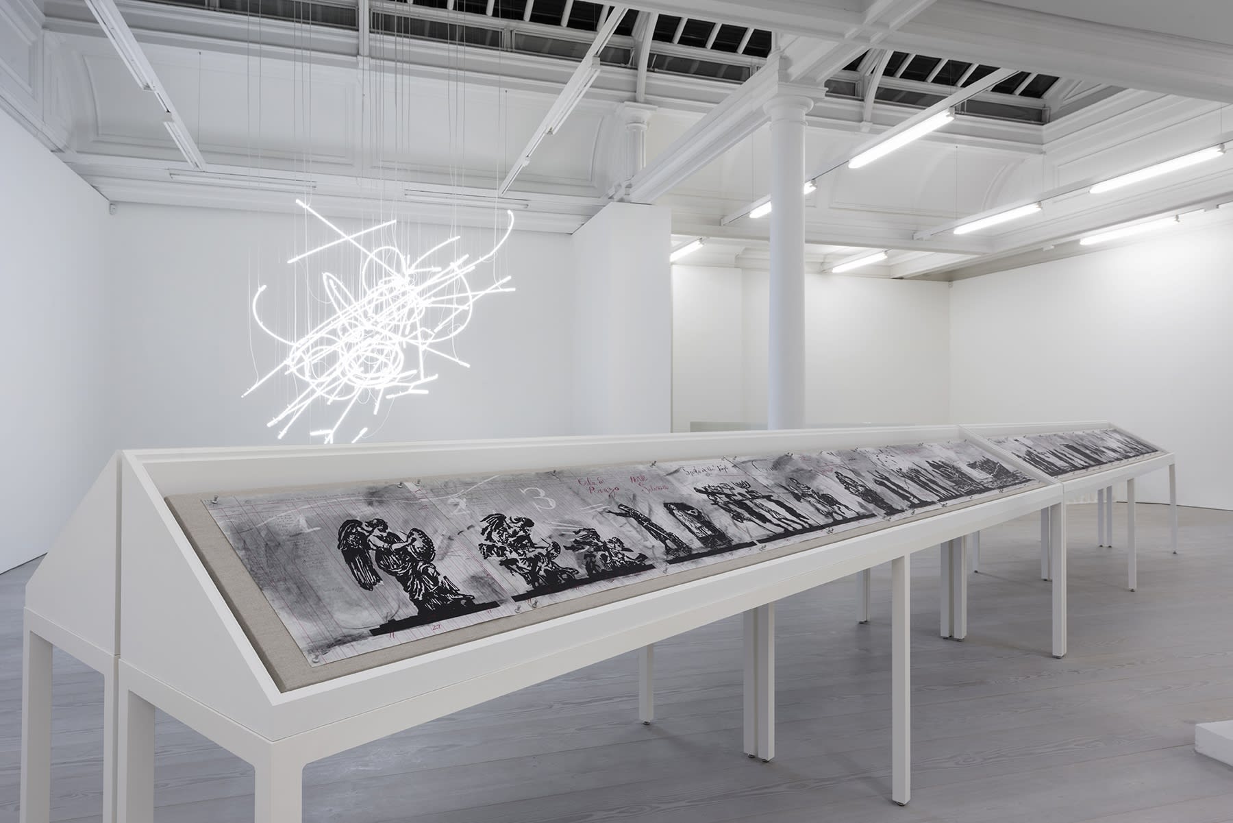 A long vitrine displaying charcoal drawings sits under a neon sculpture hanging from the ceiling.