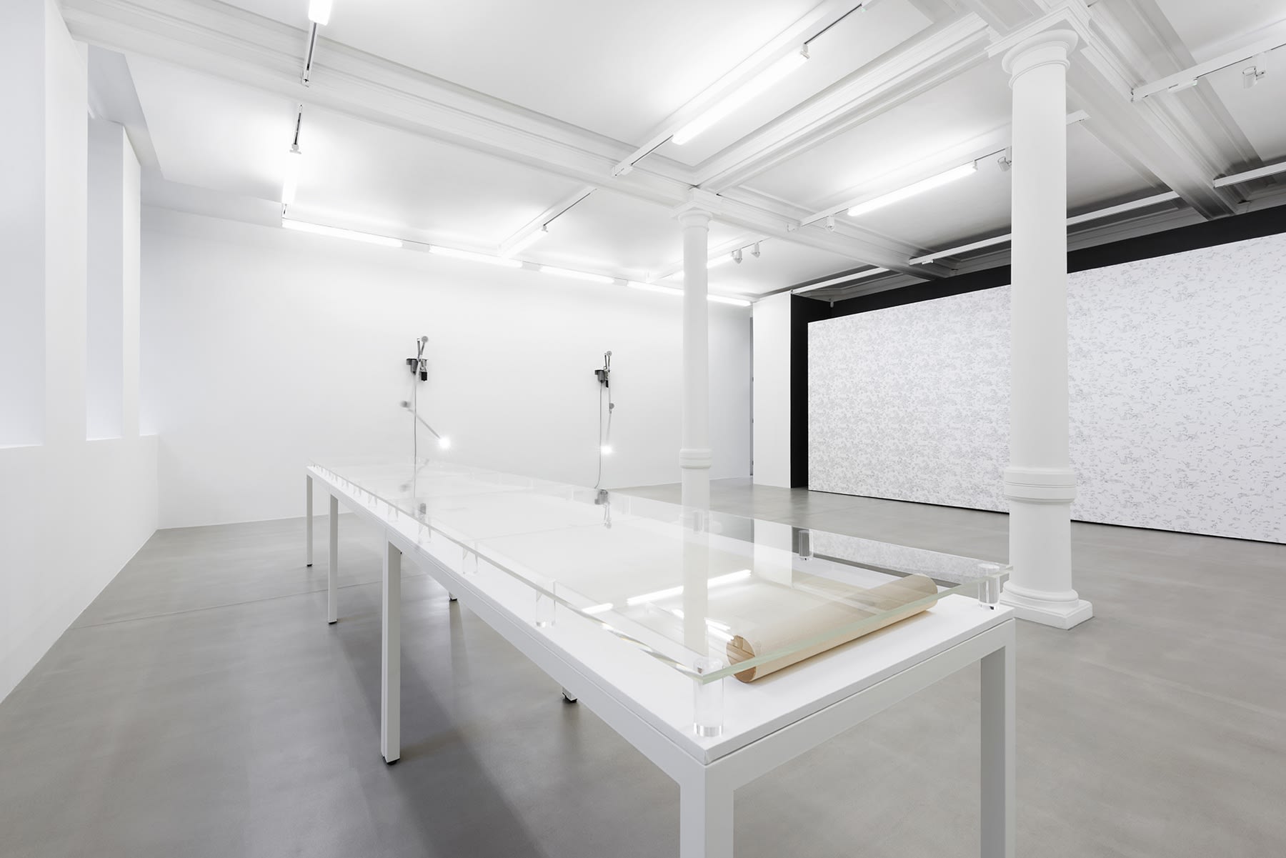 2 lights point to a long white display table in a white gallery space with columns.