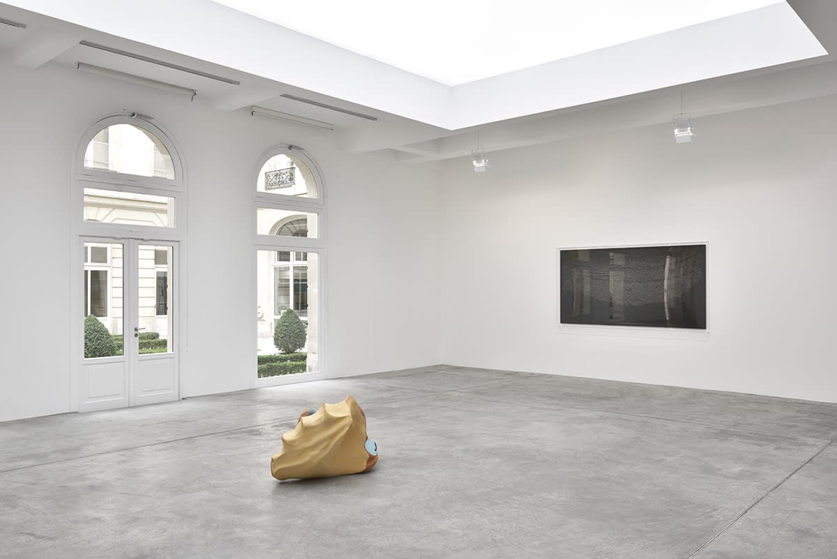 In the middle of a large white space with windows and a skylight, a wooden sculpture sits on the floor. A black painting, framed in glass, hangs on the wall in front of it.