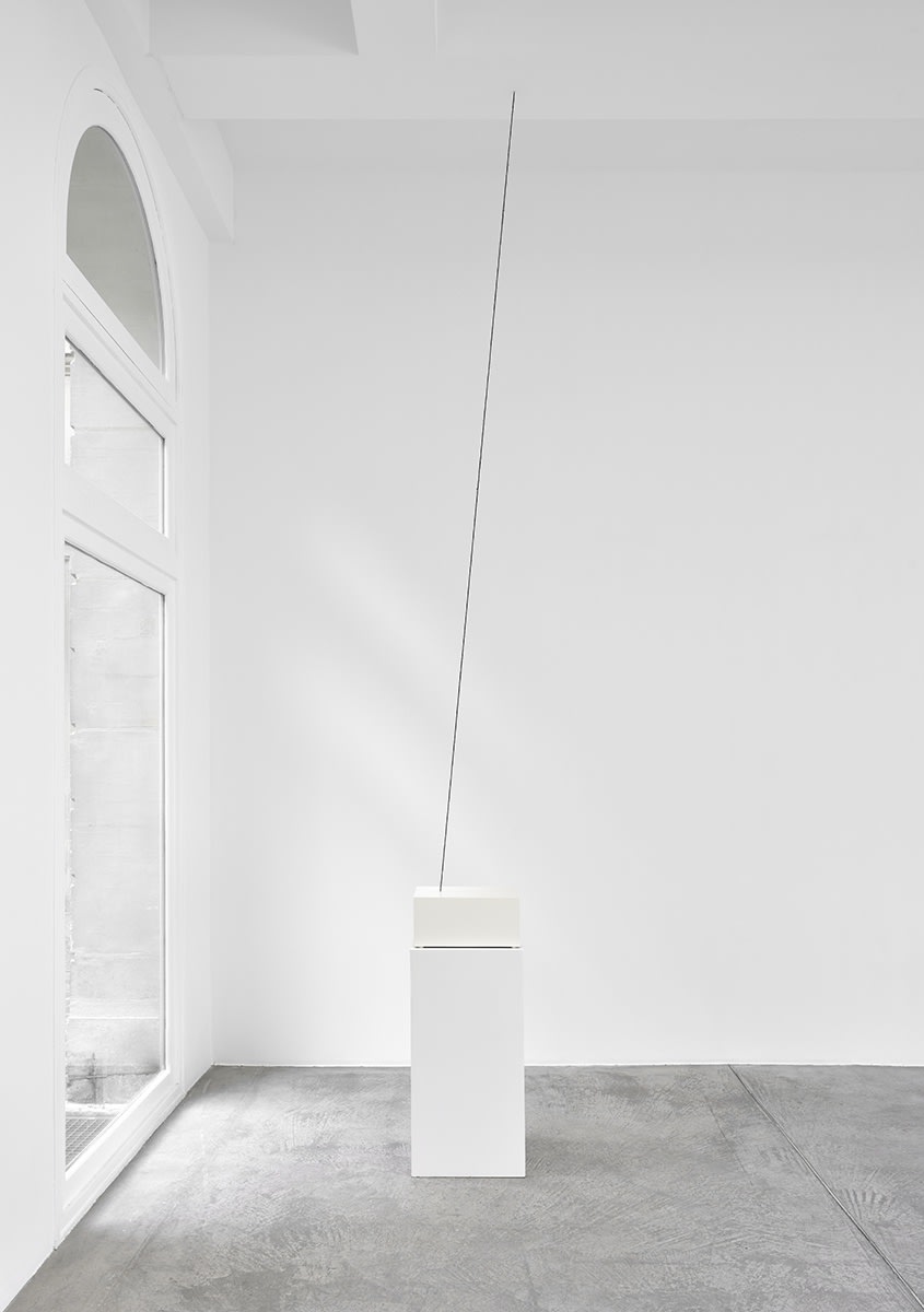 A pedestal sits in front of a window in a well lit gallery. A dark line is suspended from the ceiling.