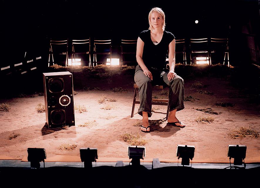 A woman sits on a chair on a stage covered with dirt and dry grass. She is well lit and stares straight ahead. 