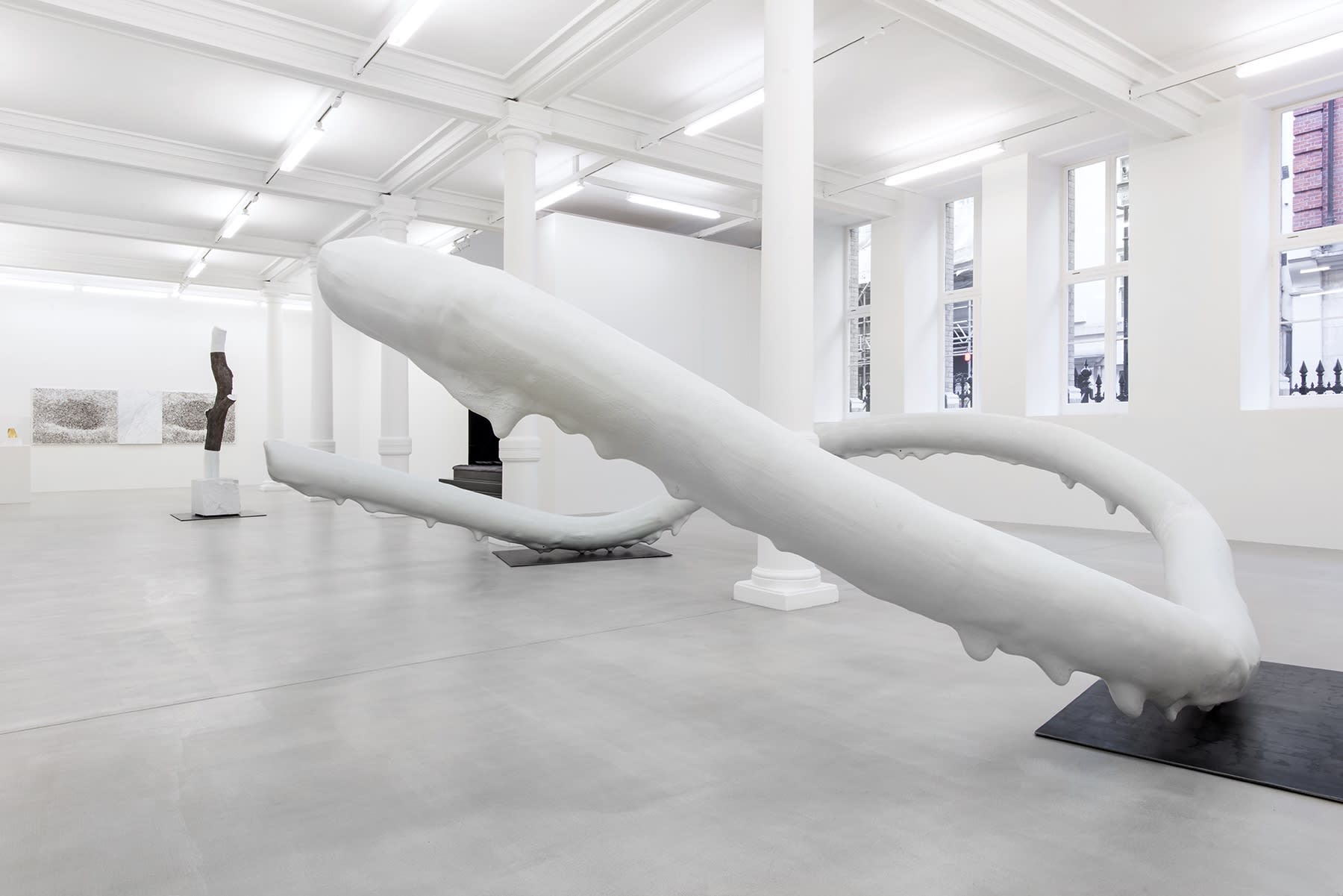 An abstract sculpture that appears to be dripping with white paint sits among other artworks in a gallery.