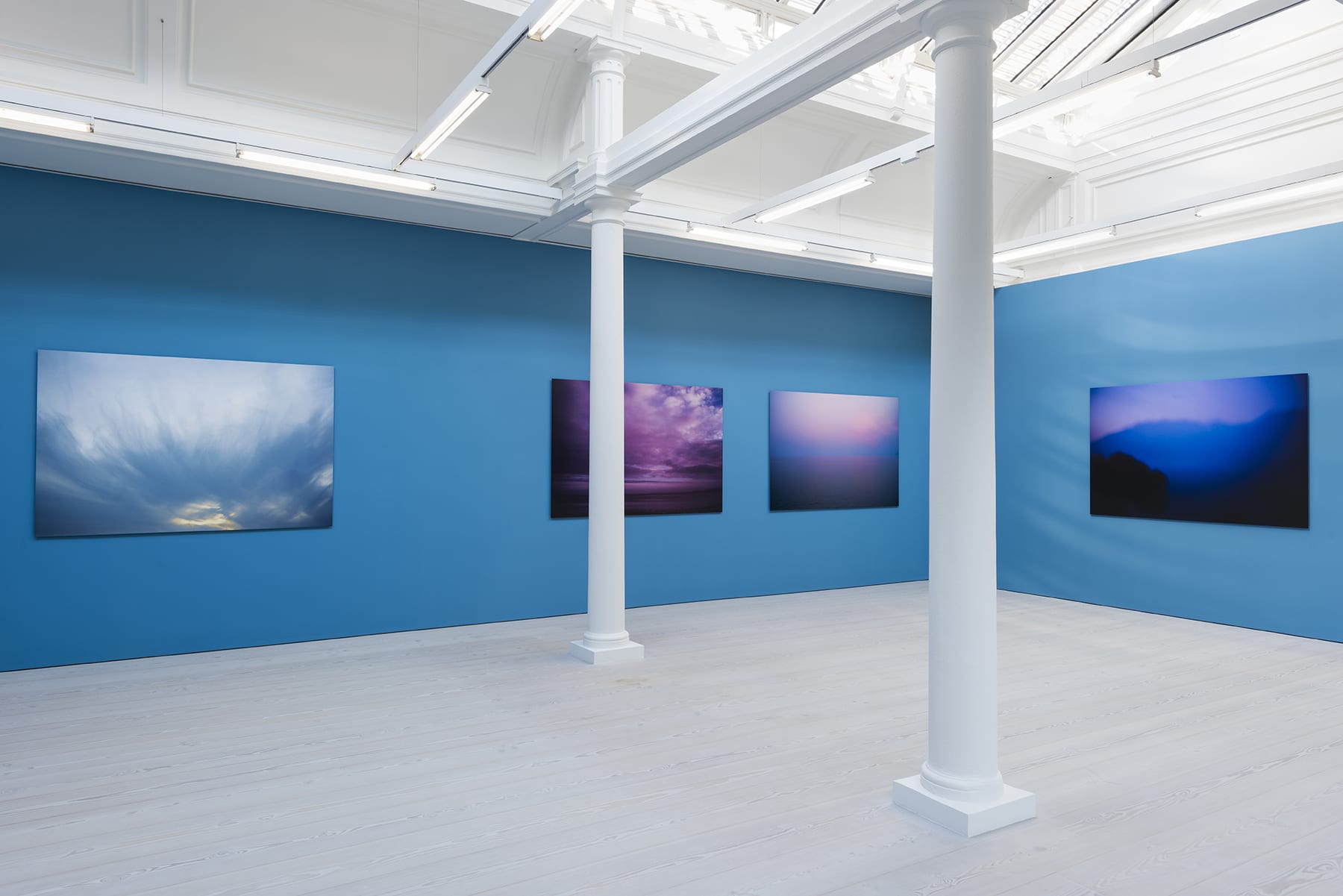 4 large color photographs of the sky hanging on a bright blue wall in a room with skylights and 2 columns.