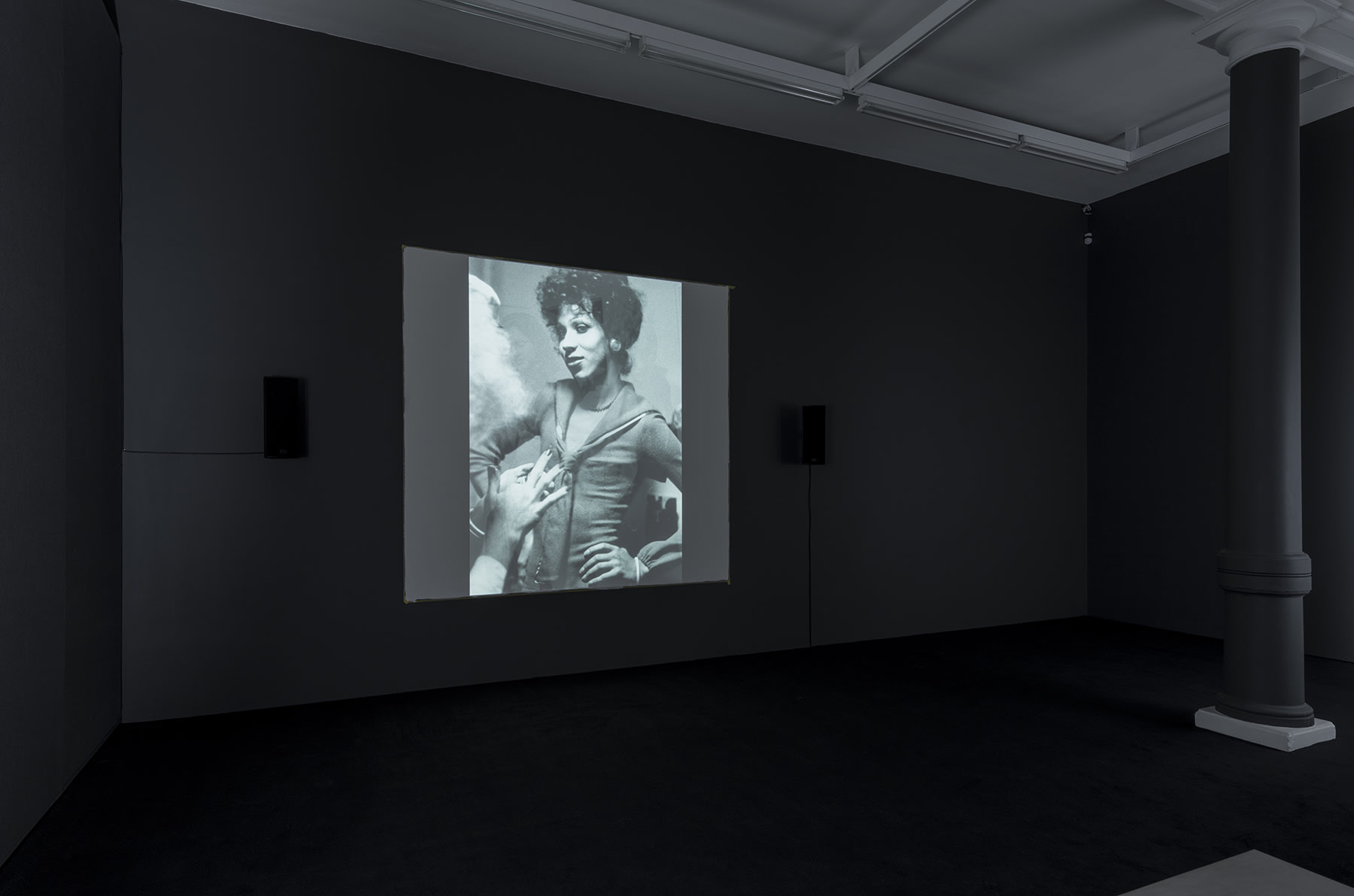 A black and white projection of 2 people talking, one with their back to the screen and the other with hands on their hips.