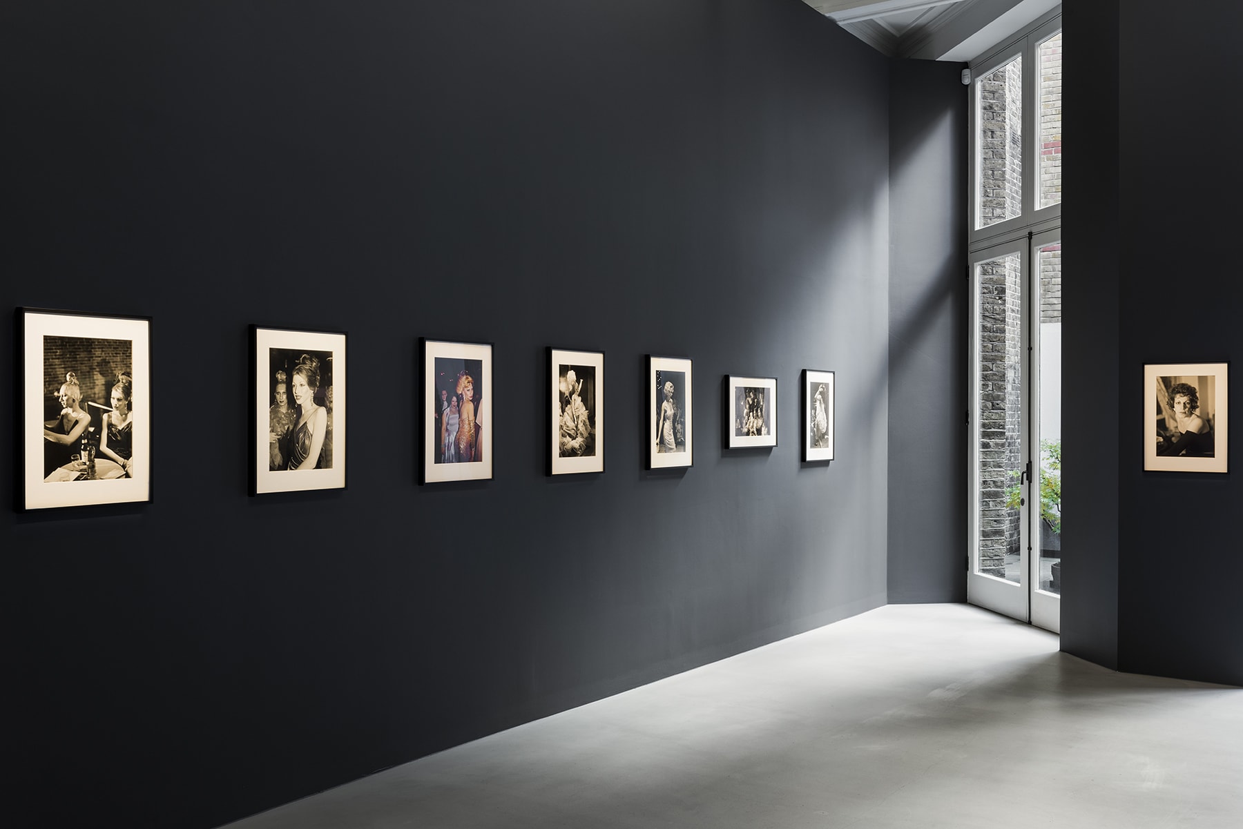 8 framed photographs hanging on a dark grey wall with a glass door in the center. 