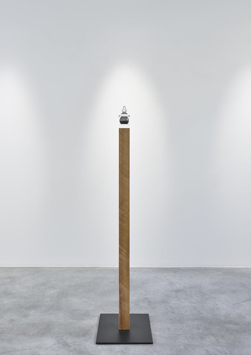 Gallery installation: singular wooden plank vertically placed with a small glass orb sculpture resting on top.