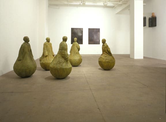 Installation view of 5 sculptures of human forms and 2 framed artworks on the wall. There is a column to the right. 