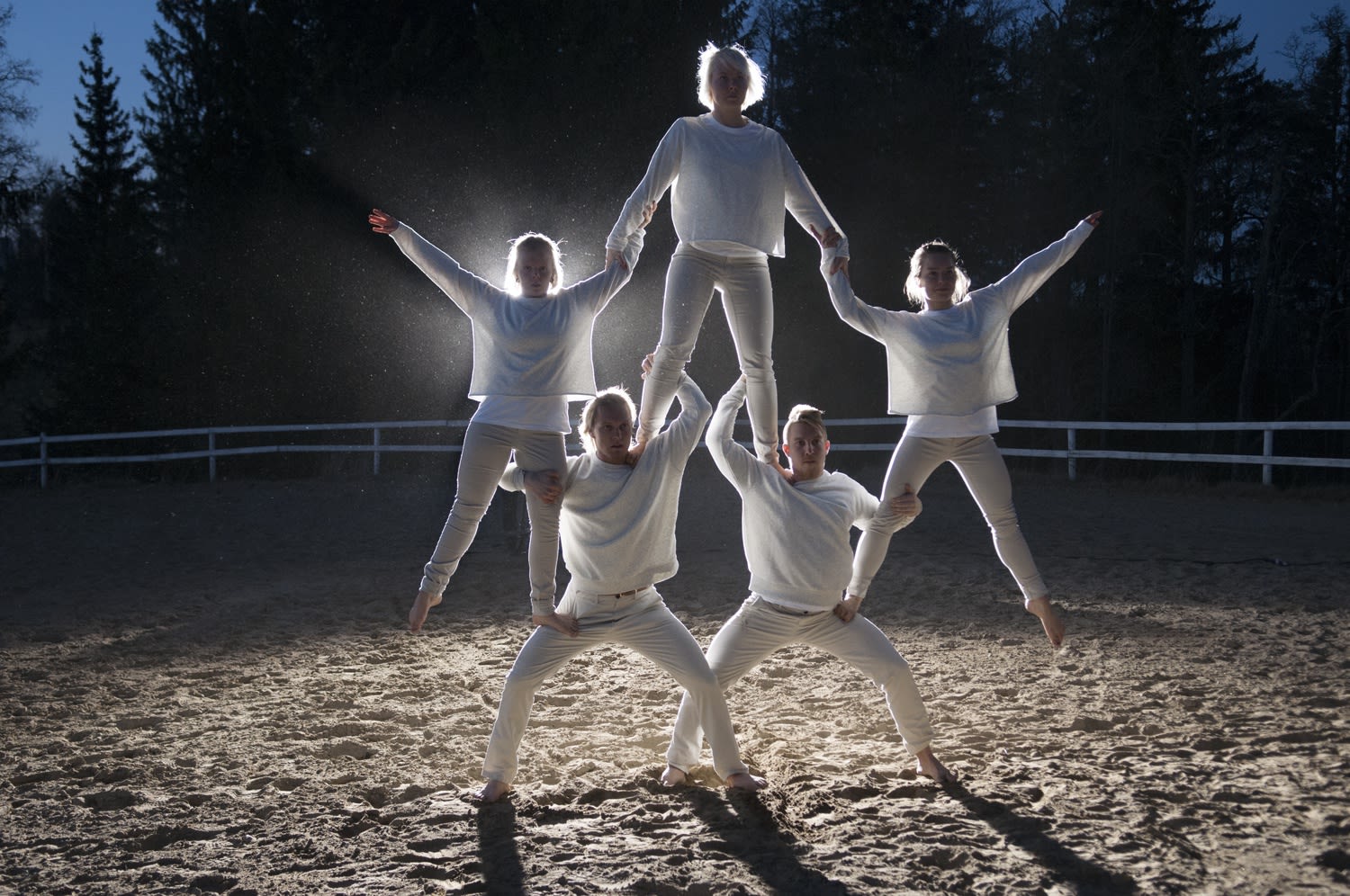 Five dancers in all white create a pyramid structure illuminated by a spotlight.