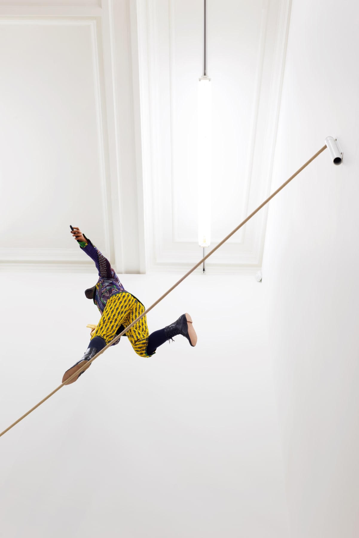 Installation view from underneath of miniature figure with a cow head and human body in wildly colored garments on a tightrope clutching a gun.