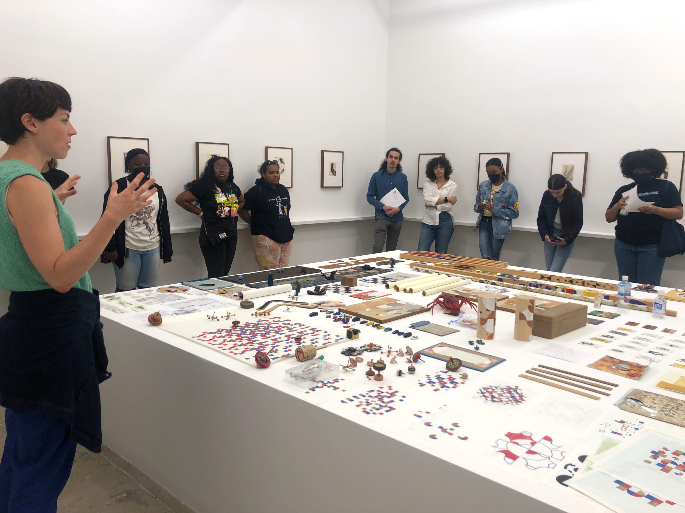 A group of students visit an exhibition by Gabriel Orozco and stand around assorted objects on a large table
