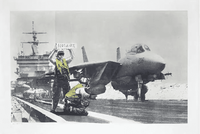 A man holds up a sign asking for applause in front of a fighter jet in this provocative Banksy anti war art, ‘Applause’.