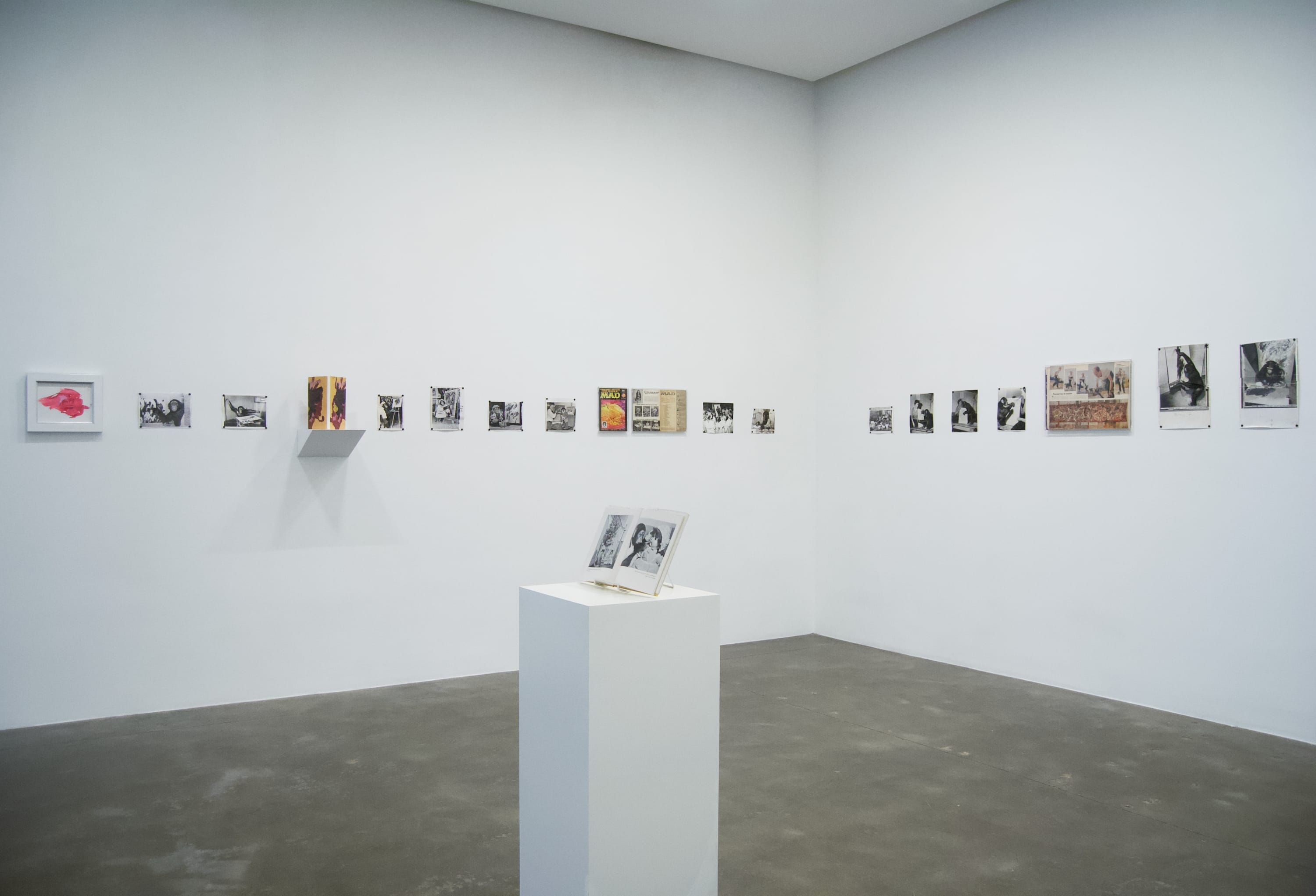 Installation view of many archival photographs hanging on white walls with a book displayed on a plinth in the center