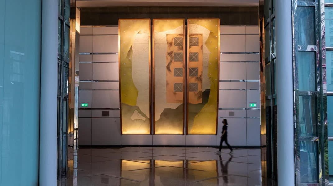 A person walks beside a large dichroic glass sculpture by Warren Carther rises from a metal wall in a corporate lobby in Hong Kong