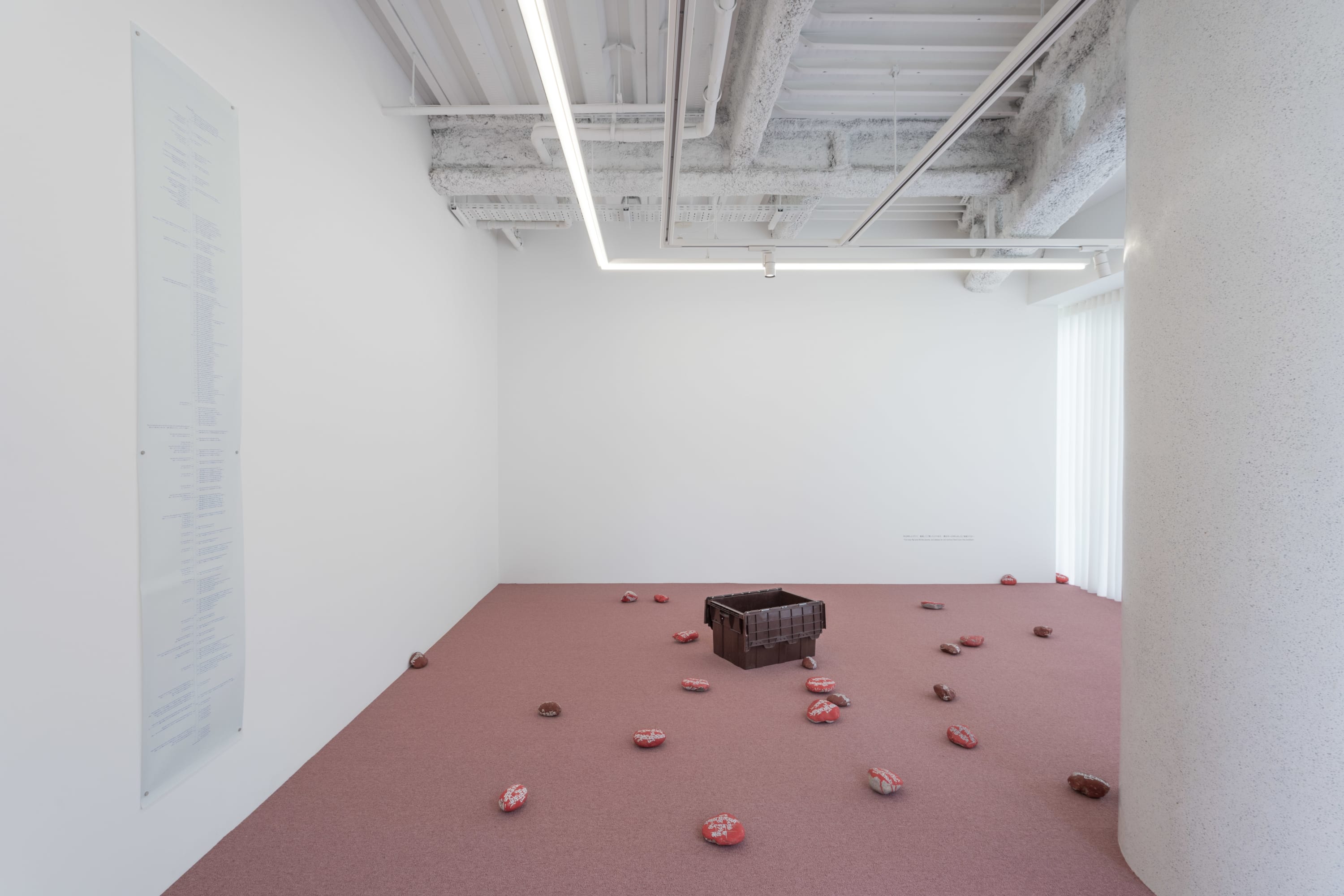 7CHOMEにおける作品は、2017年に始まり、現在も継続している長期的なプロジェクトです。ジャオ・ヤオはこのシリーズを今後も時間をかけて続けていく意志を示しています。作品の起点となっているのは、チベット地域に古くから伝わる「マニ石(マニ・ストーン)」の文化です。現地では、赤い顔料で染めた石の上にチベット仏教の経文や祈祷文を刻む職人がいて、人々は彼らに依頼して石を制作します。こうして作られたマニ石は、健康や成功などを祈願する手段であると同時に、徳を積む行為でもあります。石に経を刻むという行為は、時間と信仰を蓄積していく行為として位置づけられており、この”時間”と”信仰”という二つの概念は、ジャオの作品においても中心的な主題となっています。展示室の壁には、青焼きで印刷された石の制作タイムラインが掲げられています。制作の記録を辿ることで、“刻む”という行為が、次第に時間や日常そのものと向き合う営みへと広がっていったことを感じ取ることができます。