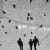 Black and white image of three shadows of figures walking among pigeons on concrete.
