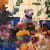 Painting of white and brown dog sitting at a table filled with fruit, flowers, cheese, bottles and glasses of wine.