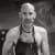 Black and white photograph of a fit, bald, shirtless man wearing an apron. He has an earring in each ear and has an intense look on his face. Art is visible out-of-focus in the backgroun.