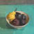 Sarah Spackman still life painting of a grey fruit bowl, with a pear and plums, sitting on a rich teal table and a grey background