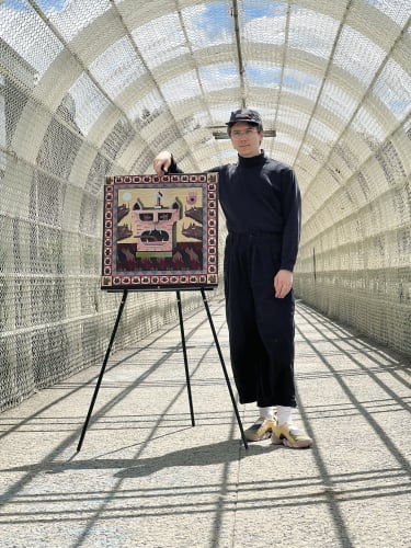 Photograph of Keya Tama Standing in a tunnel next to a painting on a black easel.