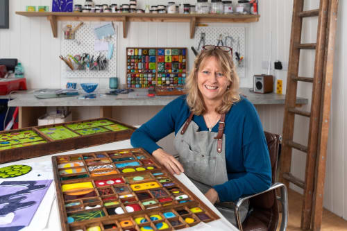 Alison Jardine in her art studio, surrounded by colourful glass artwork and creative materials.