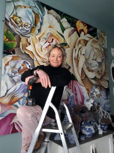 Woman seated on a ladder holding a drill, with a large floral painting and decorative vases in the background.