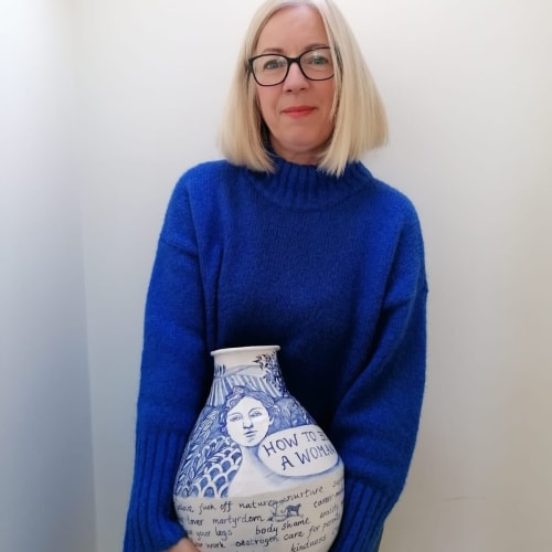 A woman in a blue sweater holding a decorative vase with blue illustrations and the text "HOW TO BE A WOMAN.