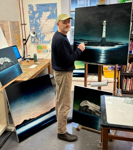 Studio shot of an artist working on a painting of a lighthouse on a rocky coast, surrounded by other maritime themed artworks.