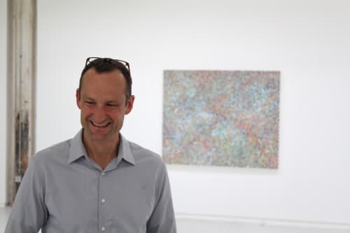 Emil Lukas standing in the gallery with small painting on wall behind him.