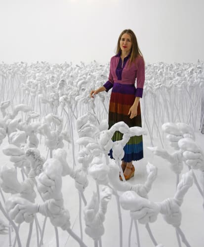 Patricia Piccinini, wearing a long rainbow striped dress stands amongst installation at Hosfelt Gallery.