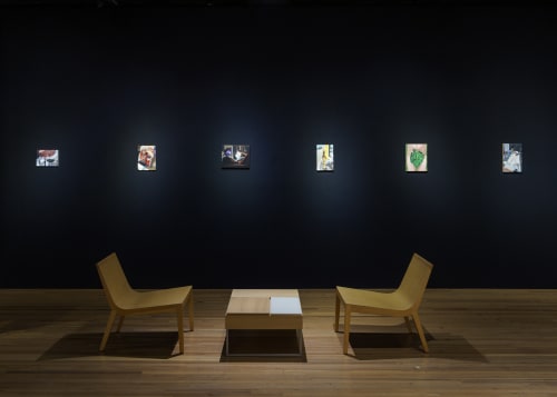 Small oil paintings in an art fair booth against a black wall illuminated by spotlight and two chairs.