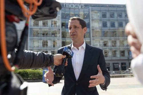 A man in a suit and white shirt is speaking to the press outdoors, gesturing with his hands as he addresses a microphone labeled “ABC 7.” The scene appears to be in front of a Macy’s storefront with reflective glass windows. The man is framed by a cameram
