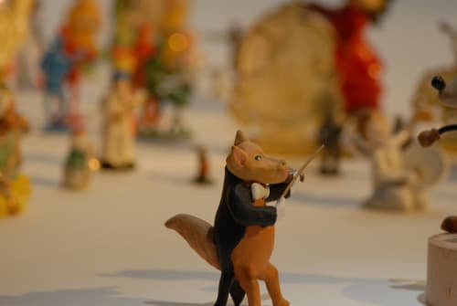 A fox in coattails plays the violin in front of of a figurine audience.